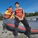 Two young boys in life vests pose with an inflatable raft on the rocky shores of a mountain river